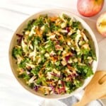 kale apple slaw in a light brown bowl next to red apples and salad servers.