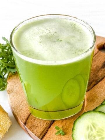 green juice in a glass on a wooden board with cucumber slices and ginger.