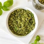 mint basil pesto in a white bowl next to fresh basil leaves and a jar of pumpkin seeds.