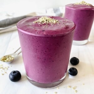 mixed berry smoothie in two glasses next to blueberries and a spoonful of hemp seeds.