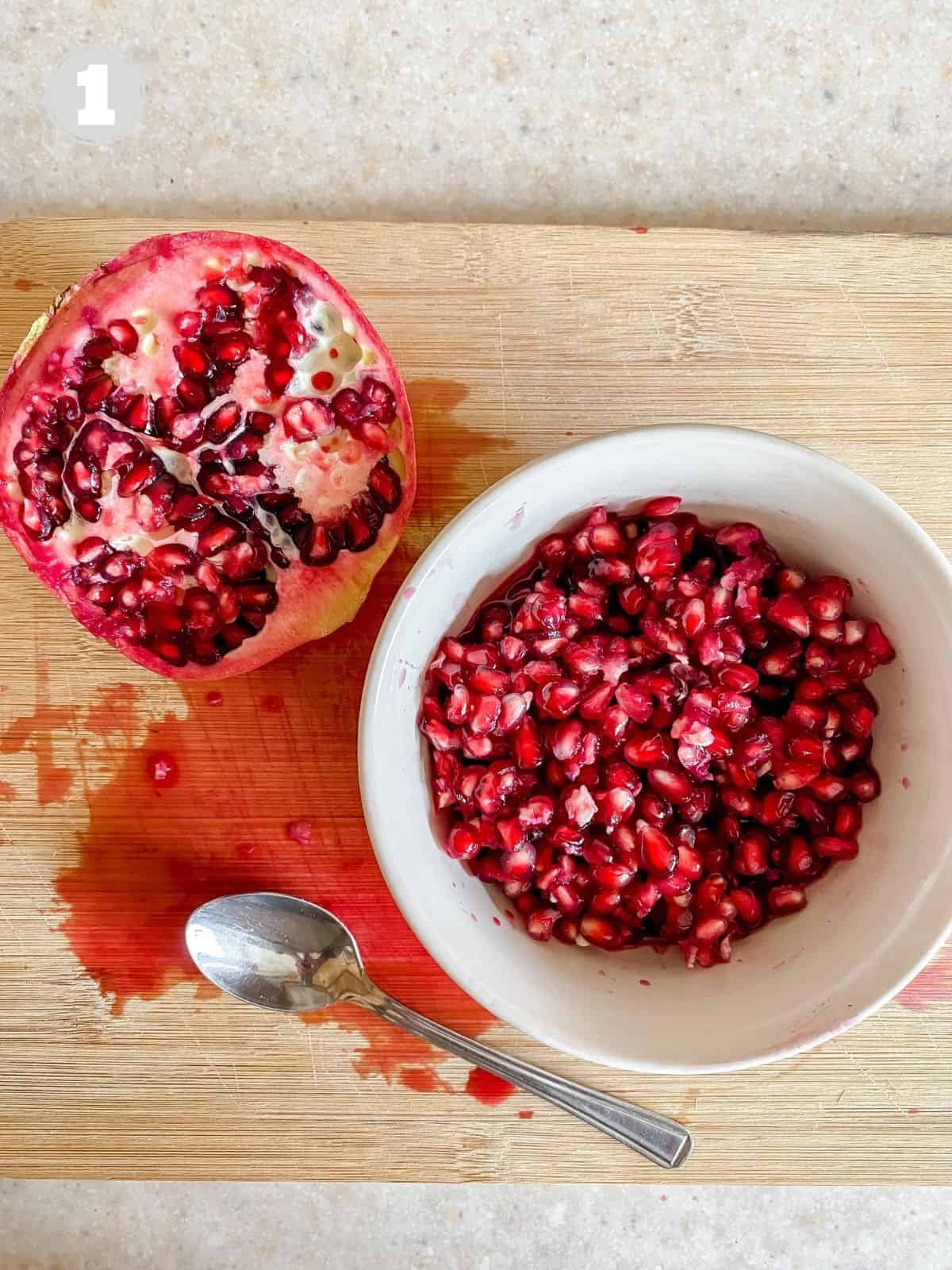 white bowl of pomegranate arils on a wooden board next to a spoon and pomegranate.