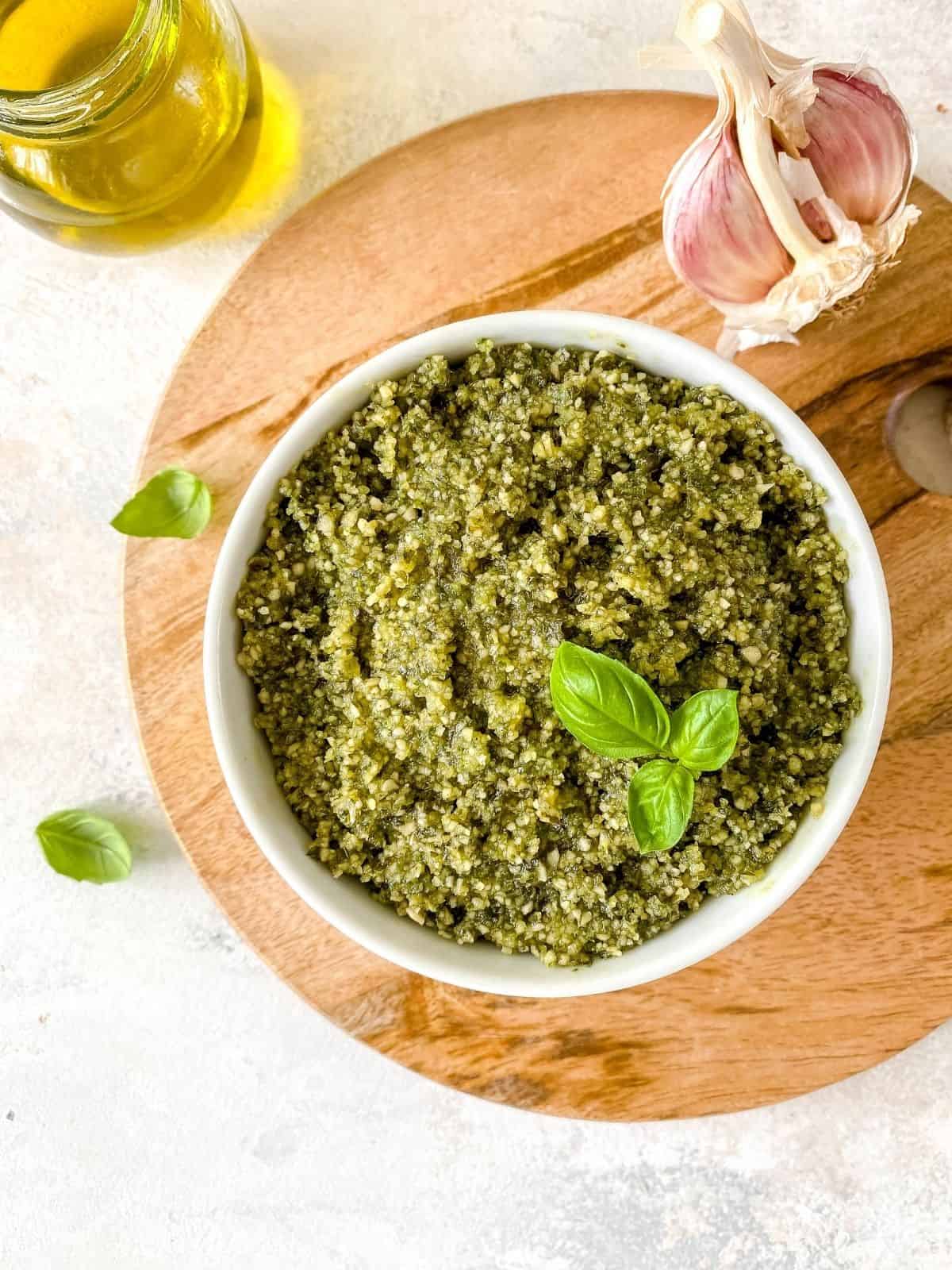 dairy free pumpkin seed pesto in a white bowl on a wooden board next to garlic and basil leaves.