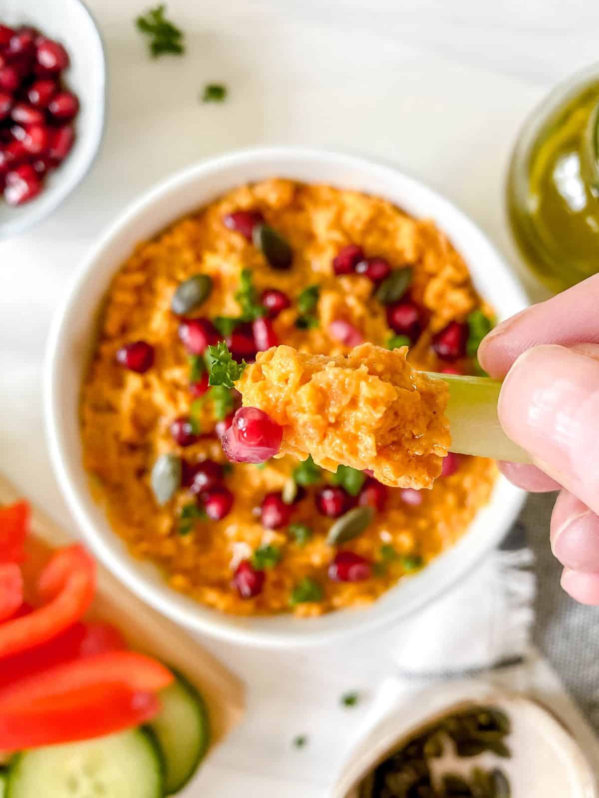 celery stick with a scoop of carrot hummus on it with a bowl in the background.