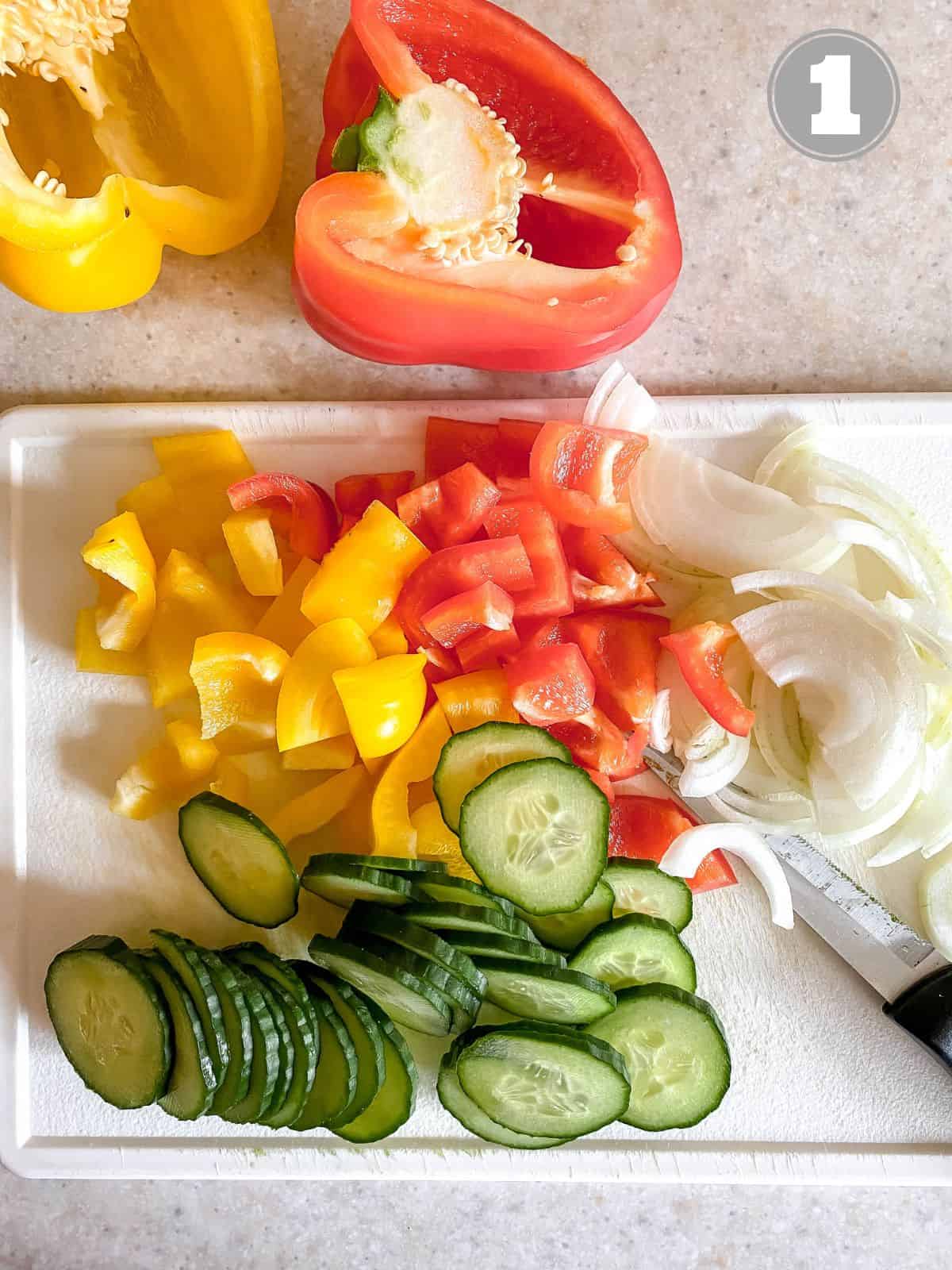 diced bell peppers, onion and cucumber on a white chopping board.