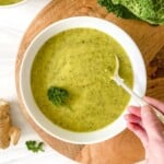 zucchini and cabbage soup in a white bowl with someone stirring it with a spoon.