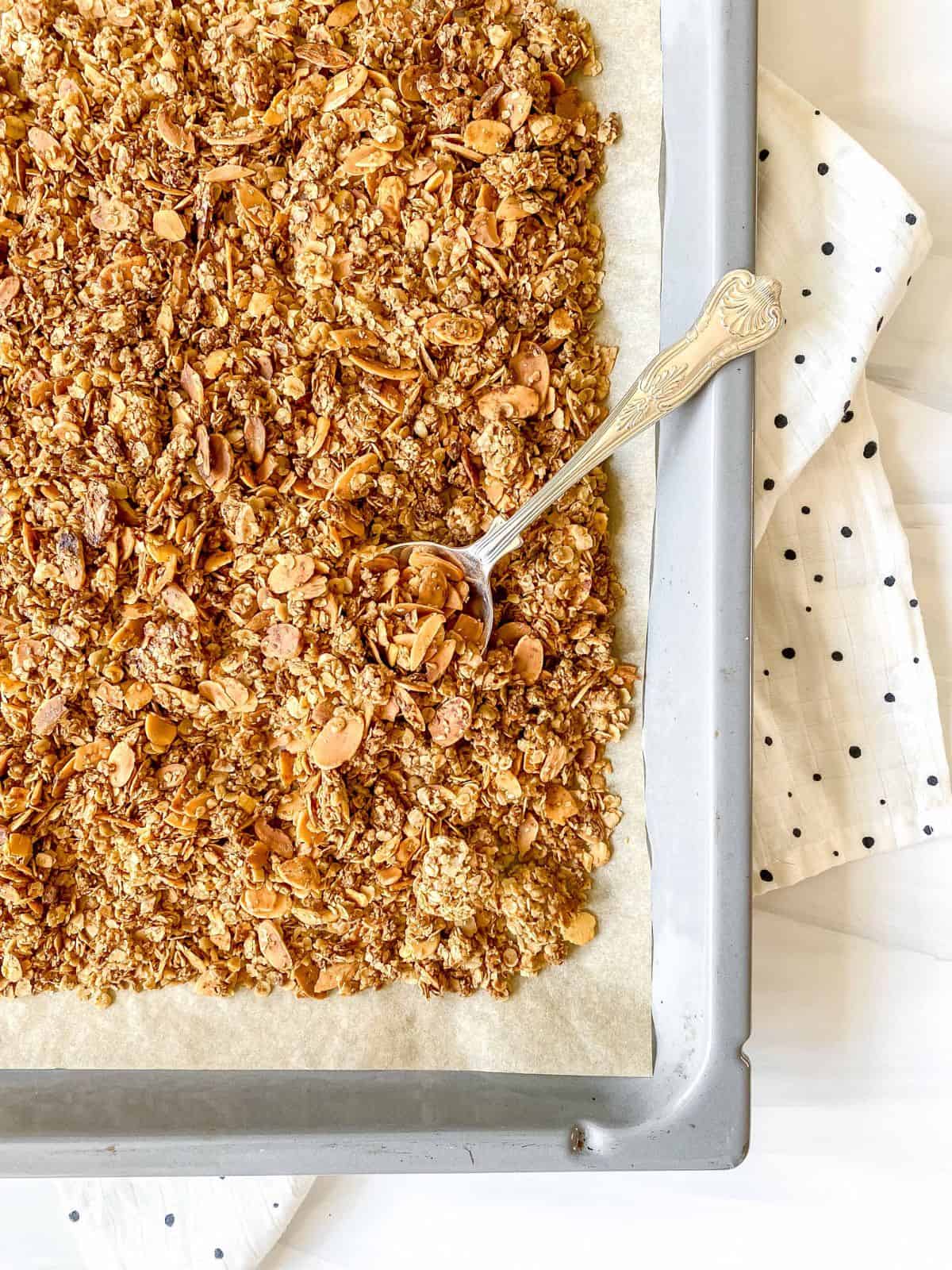 almond vanilla granola on a baking tray with a spoon in it on a spotty cloth.