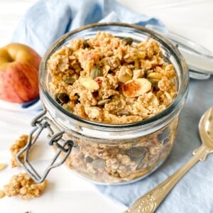 apple pie granola in a glass jar on a blue cloth with an apple.