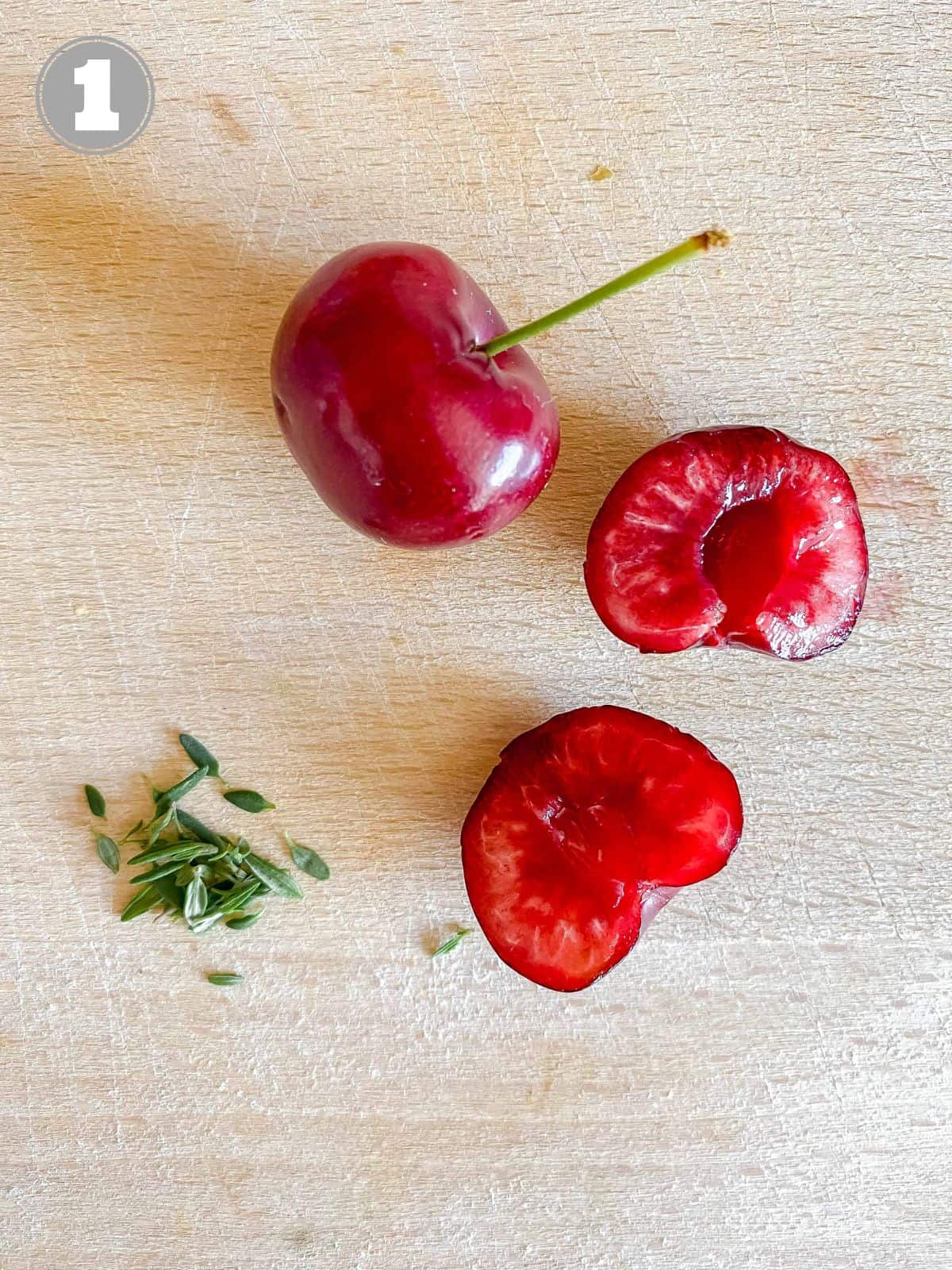 thyme leaves, a cherry and two halves of a cherry on a wooden board.