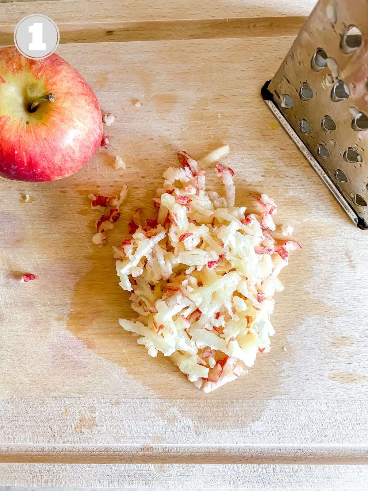 grated apple on a wooden board next to an apple and grater.
