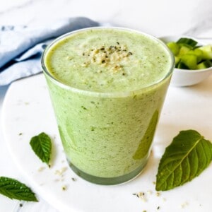 celery cucumber smoothie in a glass with a blue cloth in the background and next to mint leaves.