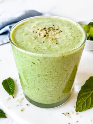 celery cucumber smoothie in a glass with a blue cloth in the background and next to mint leaves.