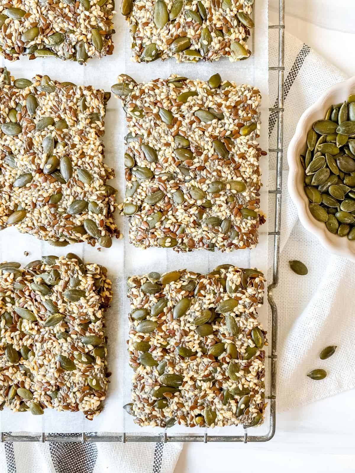 six chia and flax seed crackers on a wire rack next to a small bowl of pumpkin seeds.