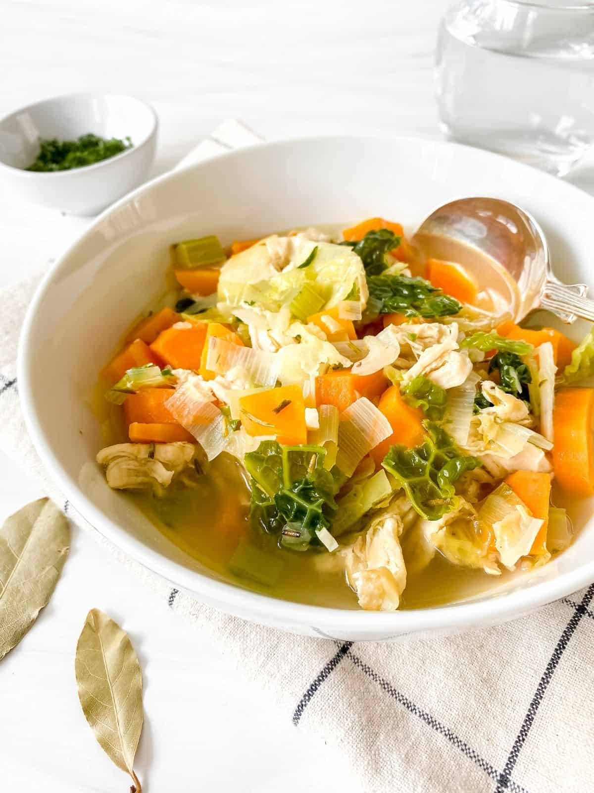 chicken and cabbage soup in a white bowl with a glass of water and bowl of herbs in the background.