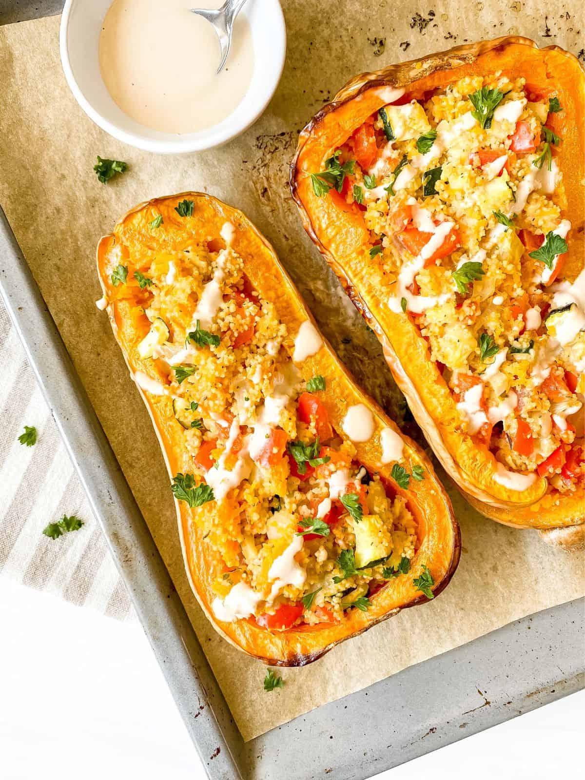 couscous stuffed butternut squash on a baking tray with a bowl of tahini.