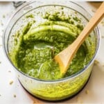 green pesto in a food processor with a wooden spoon in it.