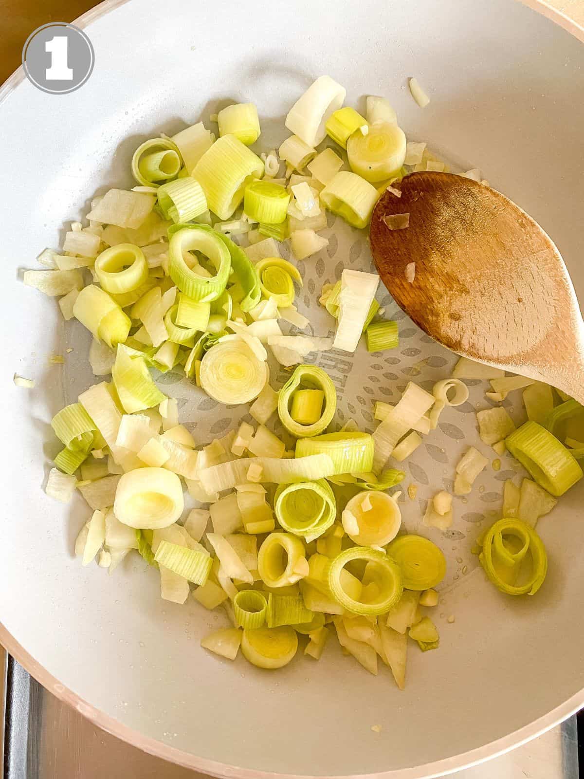 onion and leek in a light grey skillet with a spoon in it and number one.