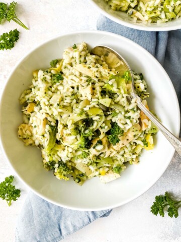 pesto chicken risotto in a white bowl with a spoon in it on a blue cloth surrounded by parsley.