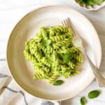 vegan broccoli pesto pasta in a light brown bowl with a fork in it next to a bowl of basil leaves.