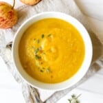 celeriac and carrot soup in a white bowl on a beige cloth next to apples.