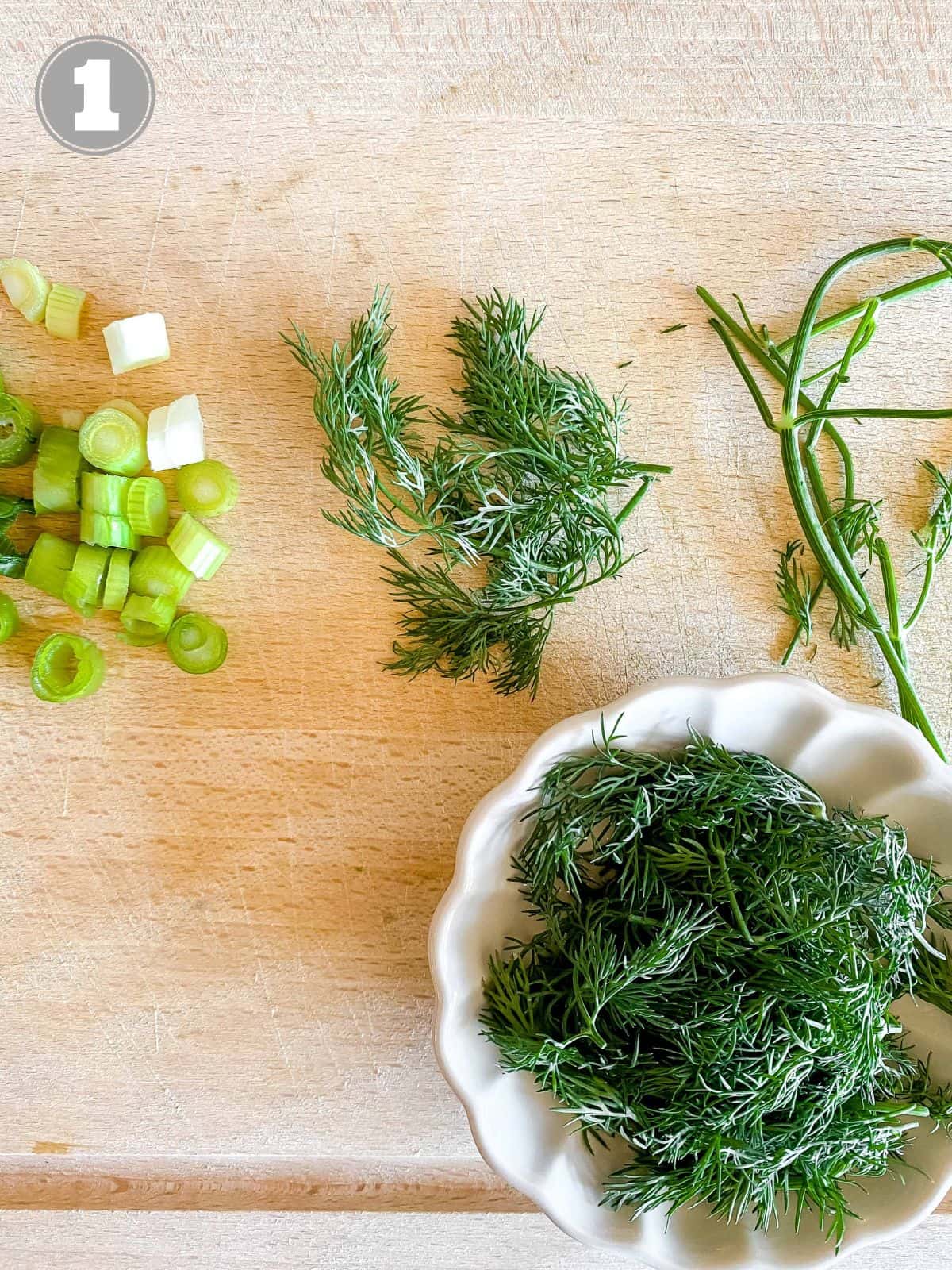 diced green onion and dill on a wooden board with dill in a bowl.