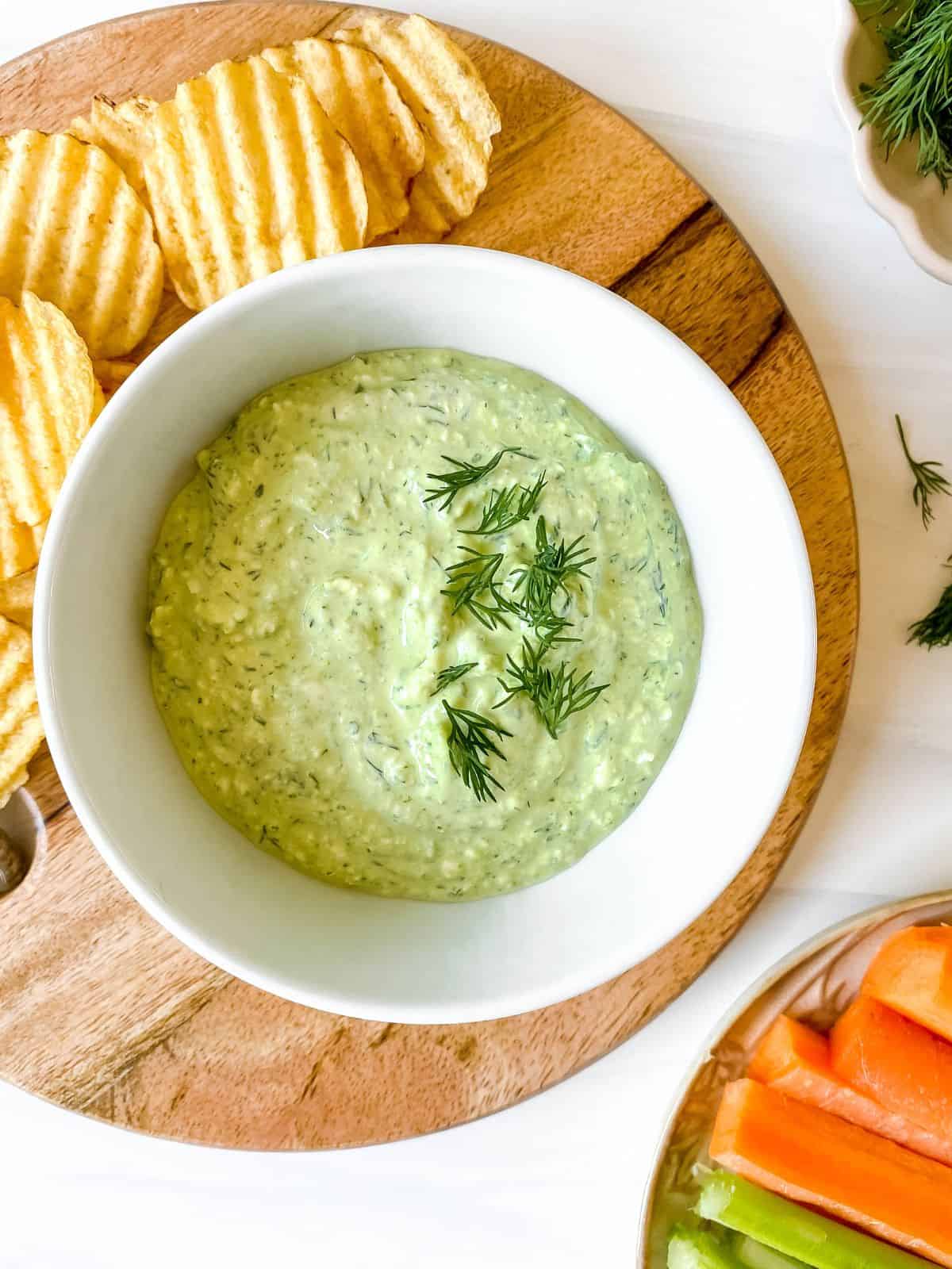 cottage cheese dill dip in a white bowl on a wooden board surrounded by crudites and chips.