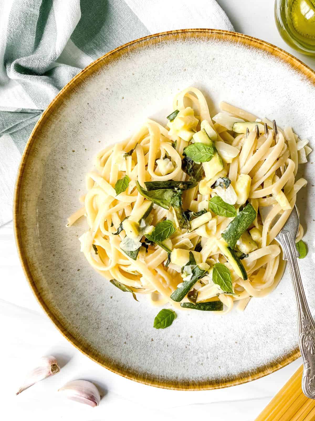 creamy zucchini pasta in a light grey bowl with a fork next to a green striped cloth and garlic.