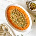 gluten free sweet potato casserole in a white dish next to a bowl of pistachios, spoon and sage leaves.