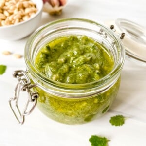 lemon balm pesto in a glass jar with a bowl of pine nuts and garlic in the background.