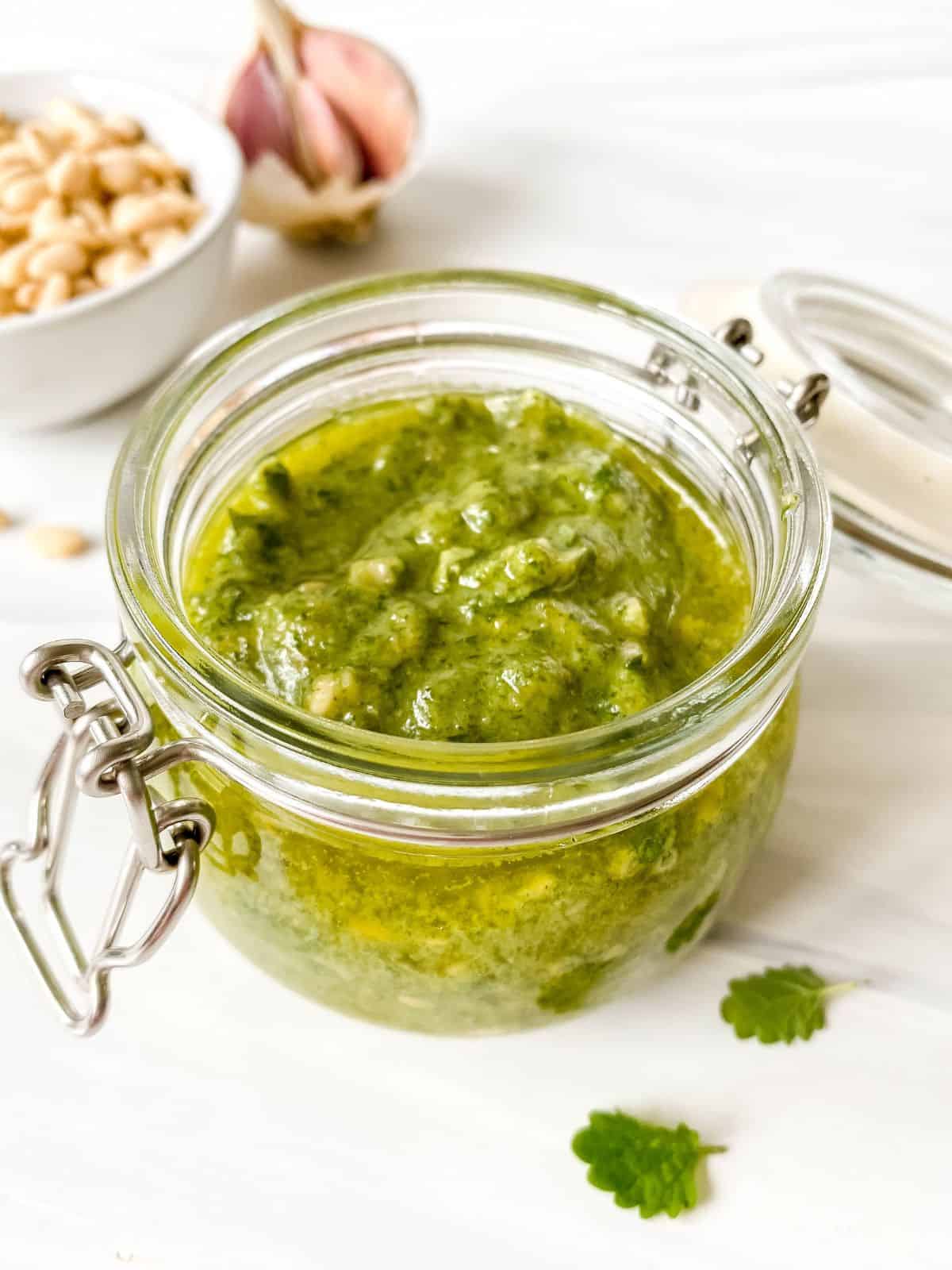 lemon balm pesto in a glass jar with a bowl of pine nuts and garlic in the background.