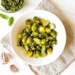 pesto Brussels sprouts in a white bowl next to a bowl of pesto, garlic cloves and basil leaves.