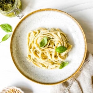 pesto ricotta pasta in a light grey bowl on a beige cloth with a for on it next to a glass jar of pesto.