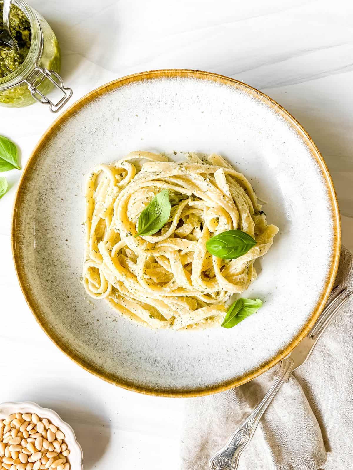 pesto ricotta pasta garnished with basil leaves in a light grey bowl with a brown rim next to bowls of pesto and pine nuts.