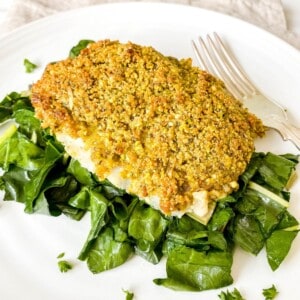 pistachio crusted cod on a bed of chard next to a fork on a white plate.