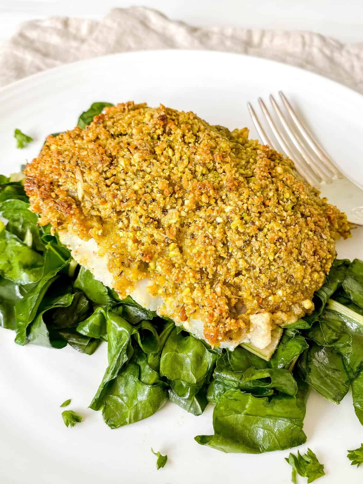 pistachio crusted cod on a bed of chard on a white plate next to a fork.
