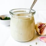 tahini dill dressing in a glass jar with a spoon it in next to garlic cloves and dill.