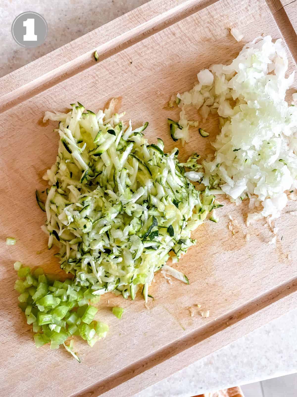 grated onion and zucchini and diced celery on a wooden board numbered one.