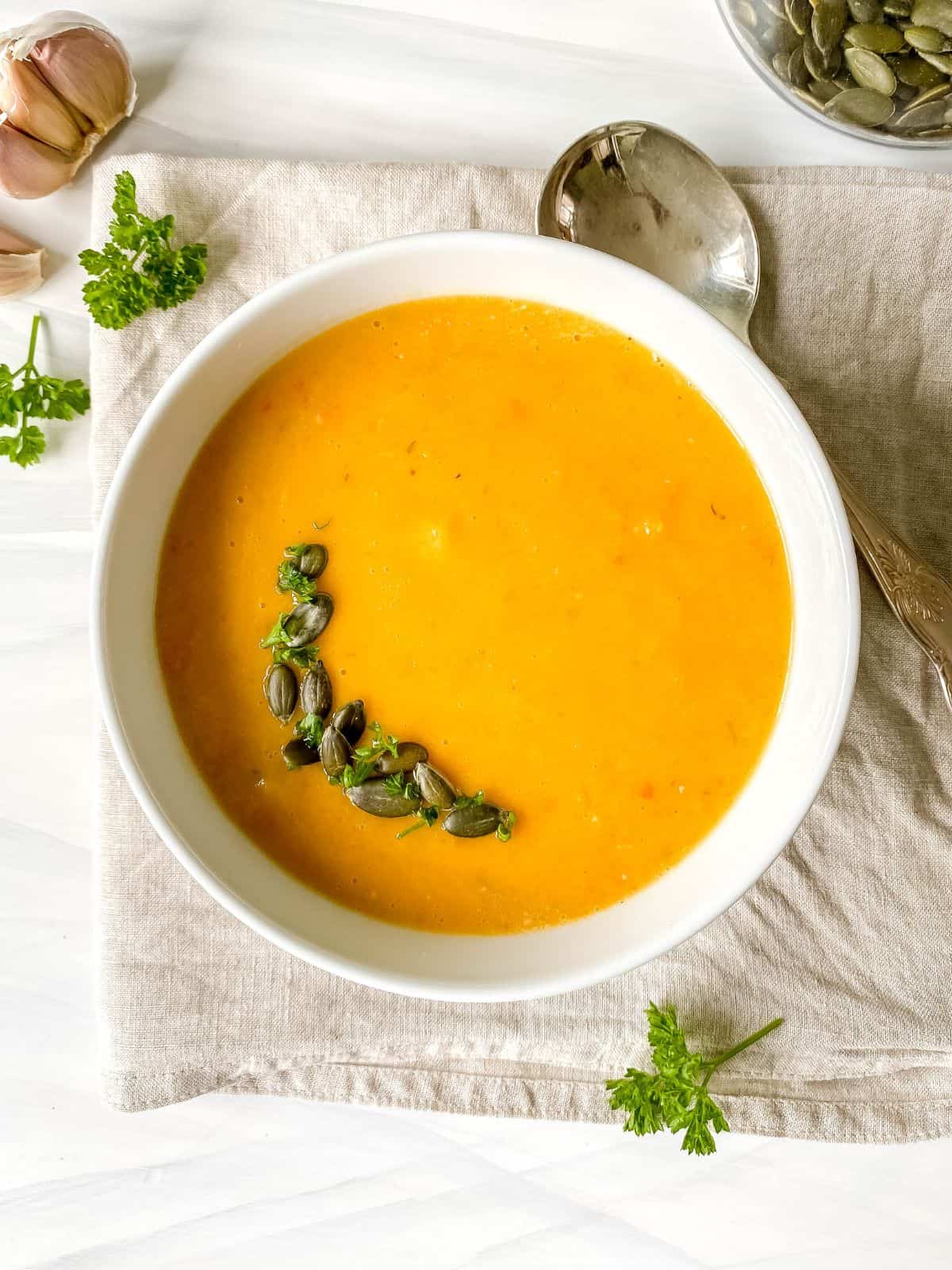 butternut squash and red pepper soup in a white bowl on a beige cloth next to a spoon, herbs and garlic cloves.
