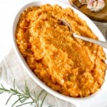 celeriac and sweet potato mash in a white dish with a spoon in next to rosemary and garlic.