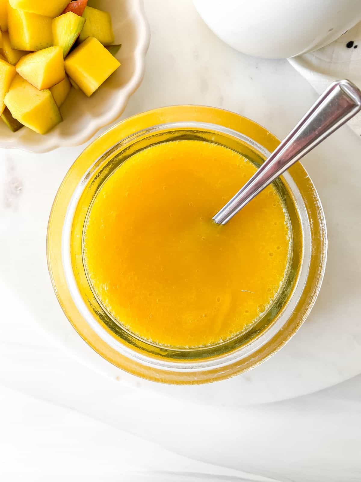 glass bowl of mango coulis with a spoon in it next to a small bowl of mango pieces.