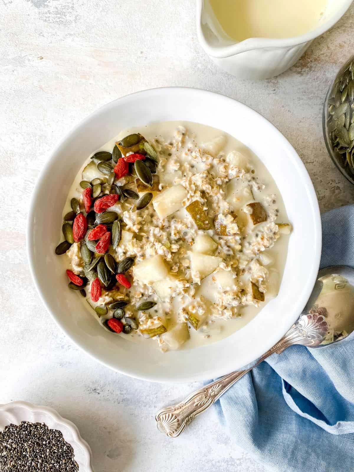 pear overnight oats in a white bowl on a blue cloth next to a jug of milk.