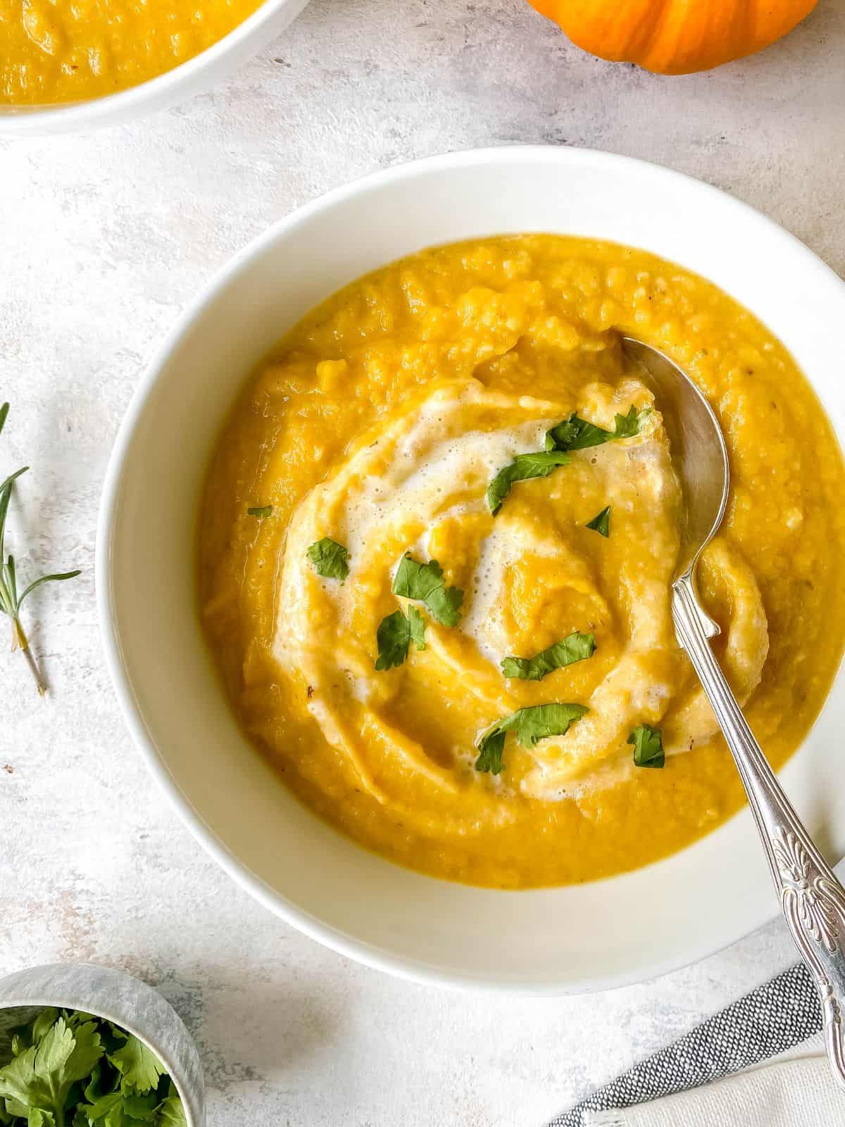 pumpkin cauliflower soup in a white bowl with a spoon in it next to a small bowl of herbs.