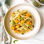 pumpkin pesto pasta on a light brown bowl next to a green striped cloth and a glass jar of pumpkin seeds.