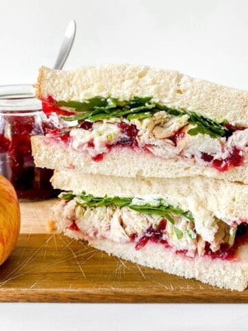 turkey cranberry cream cheese sandwich on a wooden board next to an apple with a jar of cranberry sauce in the background.