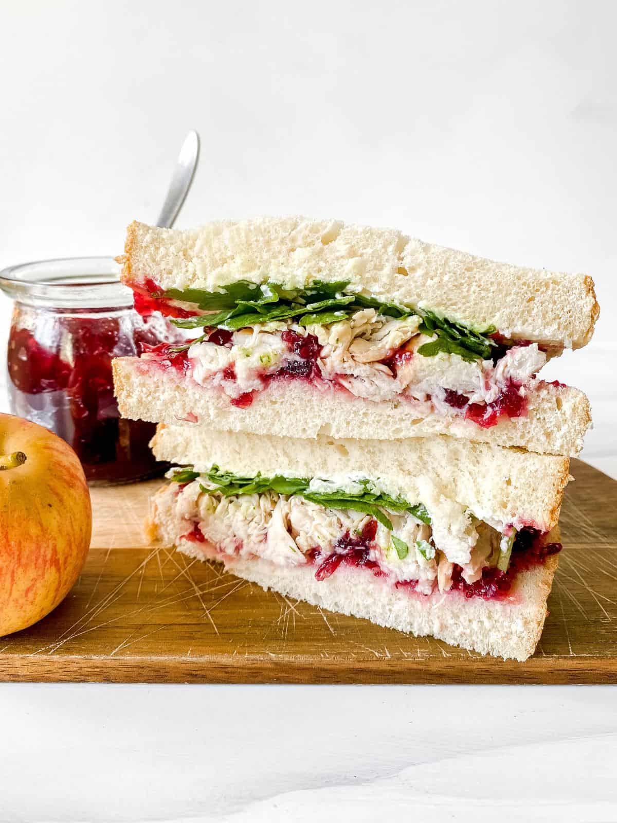 turkey cranberry cream cheese sandwich on a wooden board next to an apple and jar of cranberry sauce.