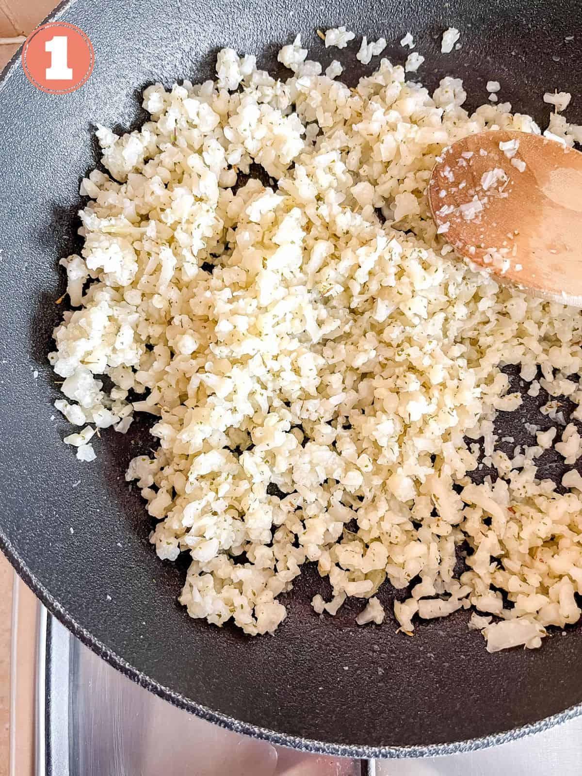 cauliflower rice in a black skillet being stirred by a wooden spoon labelled number one.