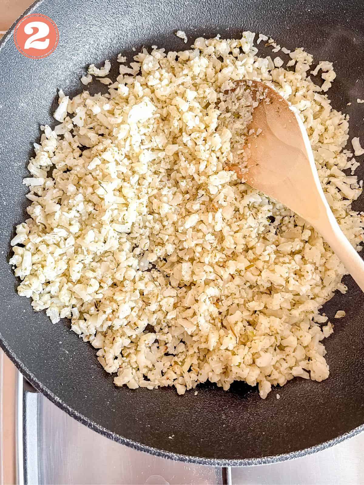 frozen cauliflower rice being stirred by a wooden spoon in a black skillet labelled number two.
