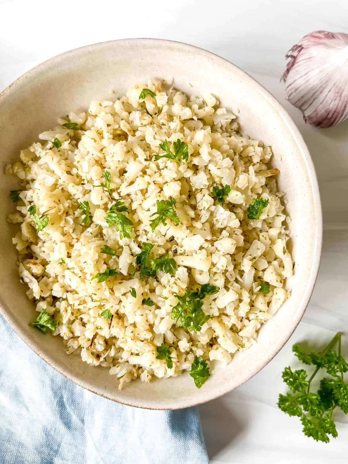 cooked frozen cauliflower rice in a cream bowl on a blue cloth next to parsley and garlic.