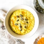 turmeric oatmeal in a cream bowl next to a glass jar of pumpkin seeds.