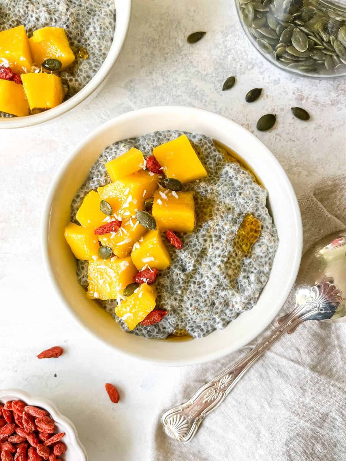 warm chia pudding topped with mango chunks in two cream bowls next to a spoon and glass jar of pumpkin seeds.