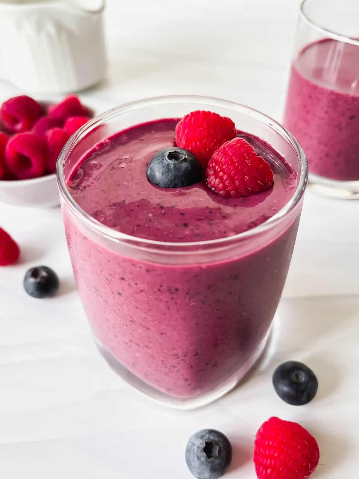 blueberry and raspberry smoothie in two glasses next to fresh fruits with a white jug in the background.